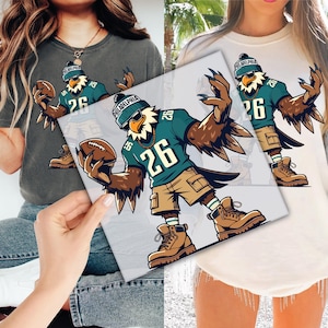 May include: A graphic design of an eagle wearing a teal jersey with the number 26, sunglasses, and a beanie that says "Philadelphia". The eagle is holding a football and wearing khaki shorts and brown boots. The design is on a t-shirt.