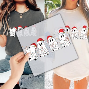 May include: A gray t-shirt and a cream-colored sweatshirt featuring a festive design of cartoon ghosts wearing Santa hats and wrapped in Christmas lights. The ghosts are holding Christmas trees and stars, creating a holiday theme.