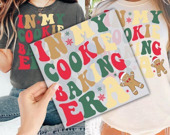 May include: Two women wearing shirts with the text "In My Cookie Baking Era" in a retro font with red, green, yellow, and brown colors. The text is surrounded by snowflakes and gingerbread cookies.