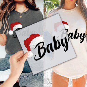 May include: Two shirts, one dark grey and one cream, each with a Santa hat graphic and the word "Baby" in a script font. The Santa hats are red with white trim and a fluffy white pom-pom.