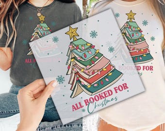 May include: A white and gray t-shirt with a Christmas tree design made of stacked books. The tree is decorated with lights and the text "All Booked For Christmas".