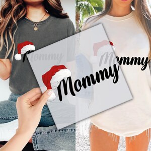 May include: Two tops, one grey t-shirt and one cream-coloured long-sleeve shirt. Both have a red and white Santa hat graphic and the word "Mommy" in black script. These are likely part of a Christmas-themed collection.
