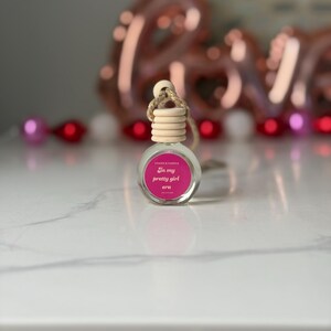 Puede incluir: Una pequeña botella de vidrio transparente con una tapa de madera y una cuerda para colgar. La botella tiene una etiqueta rosa con el texto "In my pretty girl era". El fondo incluye un globo "LOVE" borroso y cuentas decorativas.