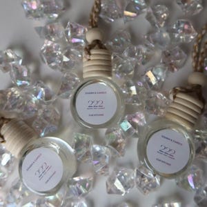 May include: Three glass car air fresheners with wooden tops and hanging strings. Each bottle has a white label with the text "Charm & Candle" and "222 CAR DIFFUSER". The bottles are surrounded by clear, iridescent crystals.