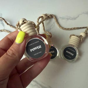 May include: Three small, clear glass car diffuser bottles with light-colored wooden caps and braided hanging cords. Each bottle features a circular black label with white text that reads CHARM & CANDLE, POWER, and CAR DIFFUSER.
