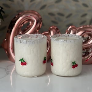 May include: Two white glass candles with a textured pattern and small red cherry accents. The candles are set against a blurred background with rose gold heart-shaped decorations. The candles are on a white surface.