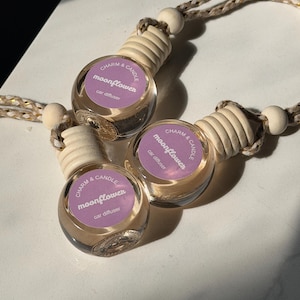 May include: Three clear glass car air fresheners with wooden tops and beige rope hangers. Each diffuser has a purple label with the words "Charm & Candle moonflower car diffuser". The bottles are arranged on a white surface.