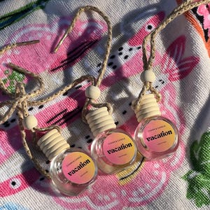 May include: Three small, round glass car air freshener bottles with light wooden caps and braided hanging cords. Each bottle features a circular label with a pink and yellow gradient background, displaying the text "CHARM & CANDLE" and "vacation".