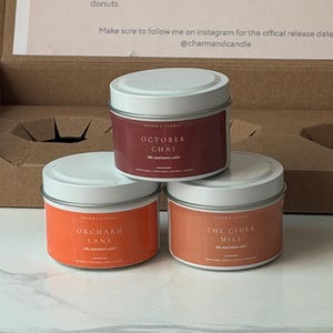 May include: Three round metal candle tins with white lids and labels in shades of red and orange. The labels feature the text October Chai, Orchard Lane, and The Cider Mill. The tins are arranged in a pyramid formation inside a cardboard box.
