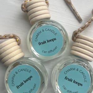 Puede incluir: Tres difusores de coche de vidrio con tapones de madera y una cuerda para colgar. Cada difusor tiene una etiqueta azul con texto blanco que dice "CHARM & CANDLE fruit loops difusor de coche".