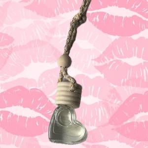 May include: Heart-shaped car air freshener with a clear glass container. The air freshener has a braided rope and a wooden bead. The background is a pattern of pink lipstick kisses.