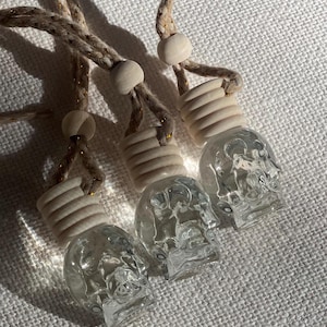 Puede incluir: Tres ambientadores para coche con forma de calavera de cristal transparente, con tapas de madera y colgantes de cuerda trenzada. Los ambientadores se muestran sobre una superficie blanca texturizada. La cuerda tiene motas doradas.