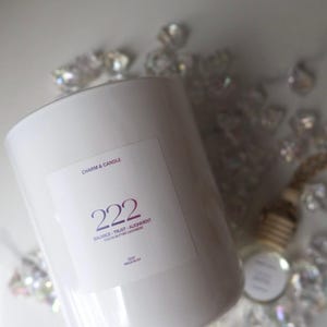 May include: A white cylindrical candle with a label that reads "Charm & Candle" and "222 Balance Trust Alignment Cocoa Butter Cashmere." The candle is surrounded by decorative crystals and a small bottle.