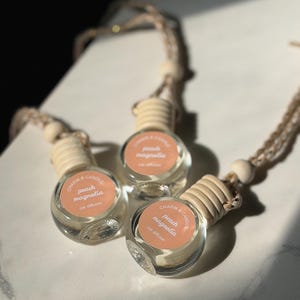 May include: Three clear glass car air fresheners with wooden tops and peach-colored labels. Each label reads "Charm & Candle, peach magnolia, car diffuser." The fresheners hang from a beige string with wooden beads.