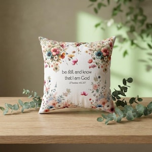 May include: Square decorative pillow with a floral design in shades of pink, blue, and yellow. The pillow features the text "be still, and know that I am God" from Psalms 46:10.