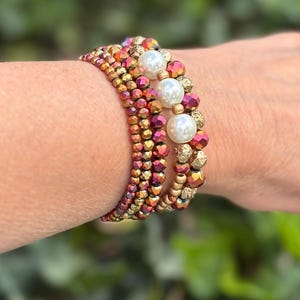 May include: A stack of beaded bracelets in shades of red, gold, and iridescent colors. The bracelets feature small, faceted beads and larger, round, white pearl-like beads. The bracelets are worn on a wrist.