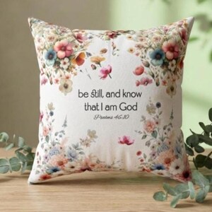 May include: A square throw pillow with a white background featuring a colorful floral border of pink, blue, and orange flowers. The center text reads, "be still, and know that I am God" followed by the citation "Psalms 46:10" in a black serif font.