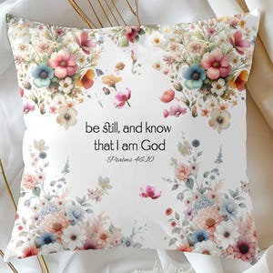 May include: Square white pillow with watercolor floral design in pink, blue, and yellow hues. The pillow features the text "be still, and know that I am God" from Psalms 46:10. Decorative home accent.