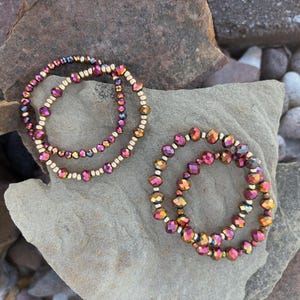 May include: Two beaded bracelets with iridescent purple, gold, and pink beads. The bracelets are resting on a gray stone surface, surrounded by other rocks. The beads are strung on a white elastic cord.
