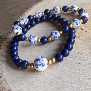 May include: Two beaded bracelets. One bracelet features dark blue lapis lazuli beads and white porcelain beads with blue floral designs. The other bracelet has similar beads. Gold-colored spacers separate the beads. The bracelets are on a light brown surface.