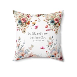 May include: Square throw pillow with a white background, adorned with floral arrangements in shades of pink, blue, and yellow. The pillow features the text "be still, and know that I am God" from Psalms 46:10.