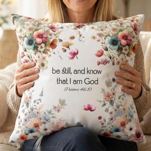 May include: A white square pillow with a floral design in shades of pink, blue, and yellow. The pillow features the text "be still, and know that I am God" from Psalms 46:10.