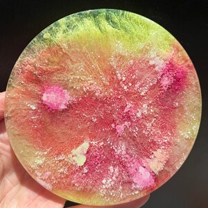 May include: A circular coaster with an abstract design. It features a mix of yellow, red, and pink colours, with white speckles and pink floral shapes. The coaster is made of resin.