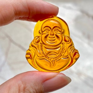 May include: A yellow glass charm depicting a smiling Buddha figure. The charm is detailed with a beaded necklace and a lotus flower design.