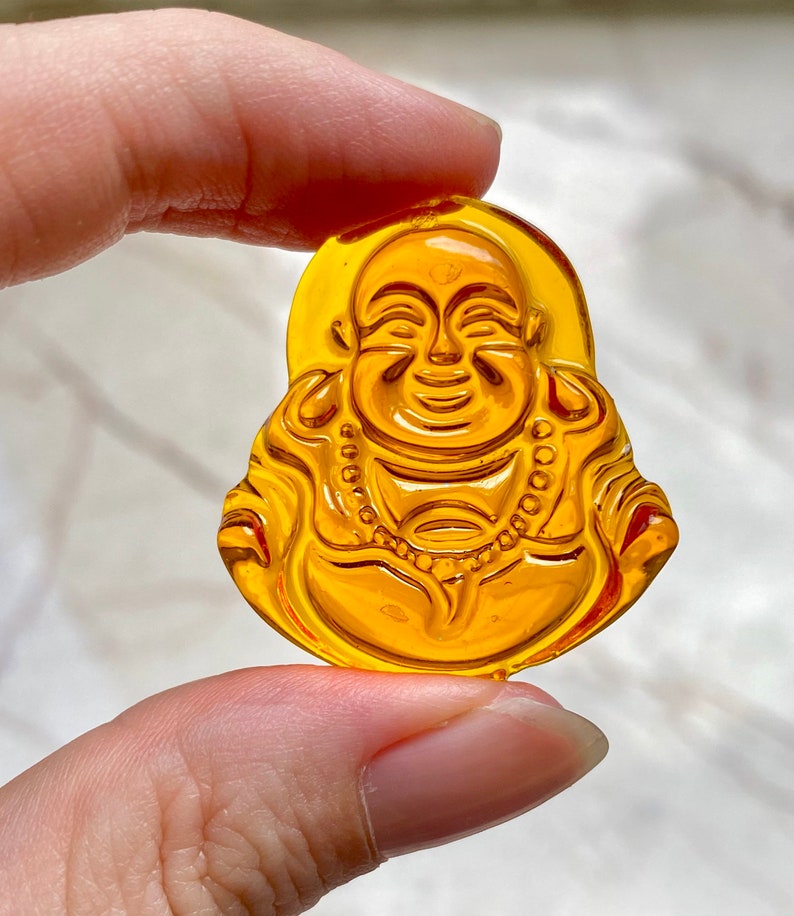 May include: A yellow glass charm depicting a smiling Buddha figure. The charm is detailed with a beaded necklace and a lotus flower design.