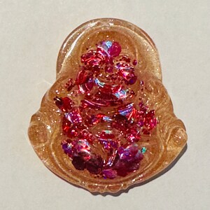 May include: A small, clear resin charm with a pink and iridescent glitter design. The charm is shaped like a seated figure with a crown and a floral design.