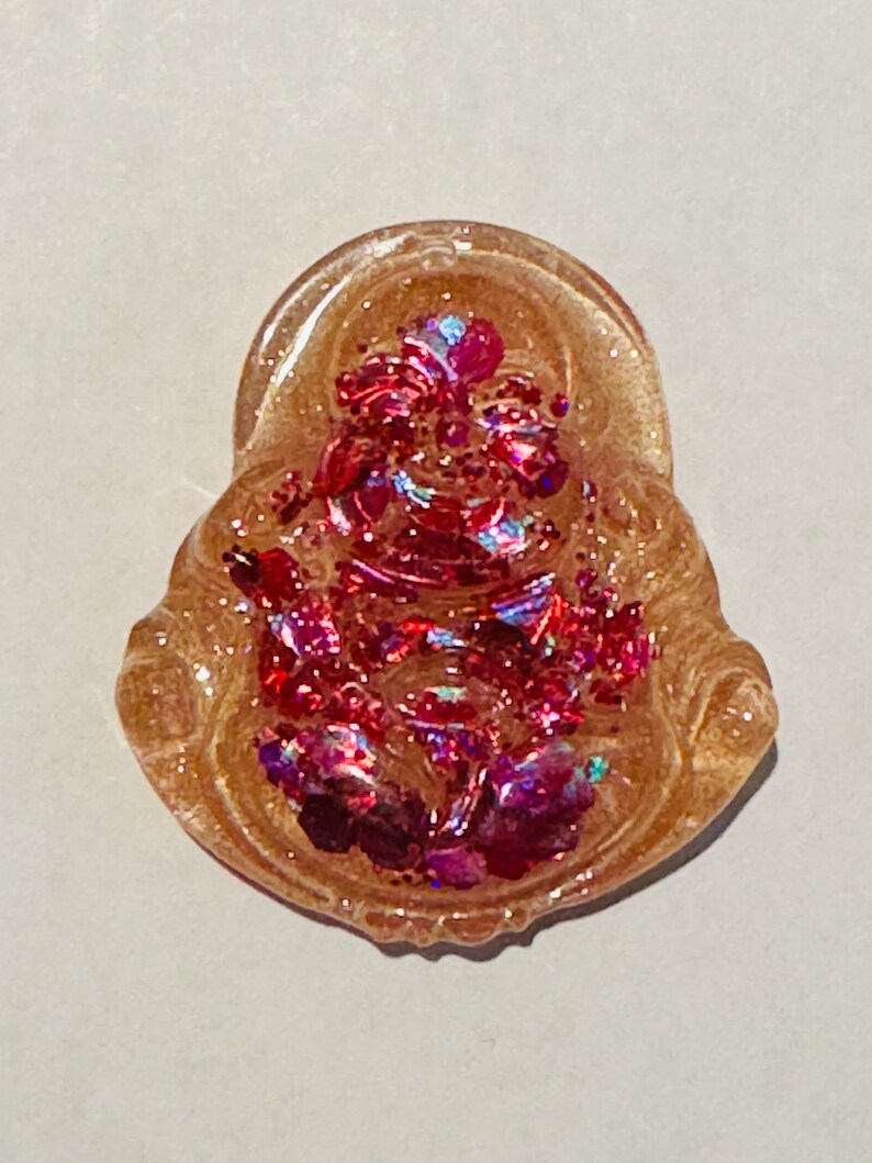 May include: A small, clear resin charm with a pink and iridescent glitter design. The charm is shaped like a seated figure with a crown and a floral design.