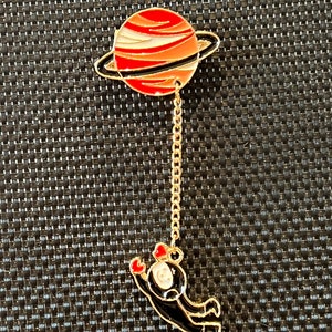 Könnte beinhalten: Eine goldfarbene Anstecknadel mit einem rot-weiß gestreiften Planeten mit einem goldenen Ring. Eine goldene Kette verbindet den Planeten mit einem schwarzen Astronauten mit roten Herzen an den Armen.