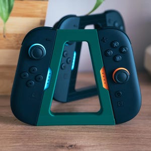 May include: Two black and green game controllers with an orange accent, resting in a green stand. The controllers have buttons and joysticks. The stand is shaped like a triangle. The background includes a wooden surface and a plant.