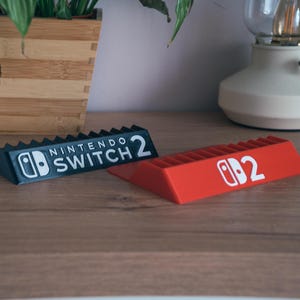 May include: Two Nintendo Switch 2 game console holders. One is black with white text and logo, the other is red with a white logo and the number 2. The holders are on a wooden surface.