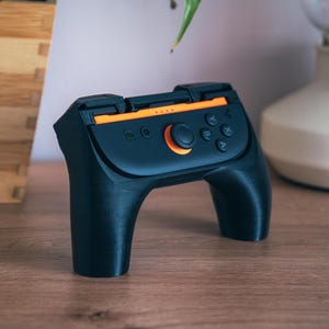 May include: Black and orange handheld gaming controller with a central orange button and several smaller buttons. The controller has a curved design with two handles and is set on a wooden surface.