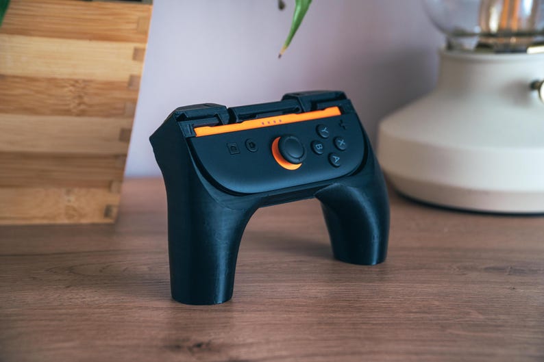 K&ouml;nnte beinhalten: Schwarzer und orangefarbener Handheld-Gaming-Controller mit einem orangefarbenen zentralen Knopf und mehreren kleineren Kn&ouml;pfen. Der Controller hat ein geschwungenes Design mit zwei Griffen, auf einer Holzoberfl&auml;che.