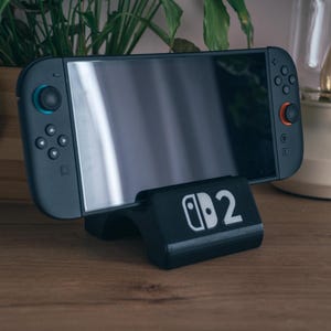 Może przedstawiać: Czarna konsola Nintendo Switch spoczywająca na czarnym stojaku z nadrukowaną cyfrą "2". Konsola ma szare kontrolery z niebieskimi i czerwonymi akcentami. Stojak stoi na drewnianej powierzchni, z roślinami i lampą w tle.