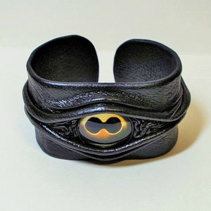 Puede incluir: Pulsera de cuero negro con un dise&ntilde;o de ojo dorado y negro. La pulsera es ajustable y tiene un dise&ntilde;o &uacute;nico y llamativo.