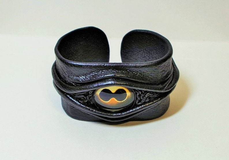 Puede incluir: Pulsera de cuero negro con un dise&ntilde;o de ojo dorado y negro. La pulsera es ajustable y tiene un dise&ntilde;o &uacute;nico y llamativo.