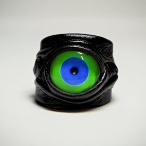 Puede incluir: Un anillo de cuero negro con un diseño de ojo verde, azul y negro.