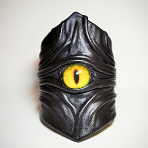 Puede incluir: Pulsera de cuero negro con un gran ojo amarillo en el centro. La pulsera está diseñada para parecerse a las escamas de una serpiente.