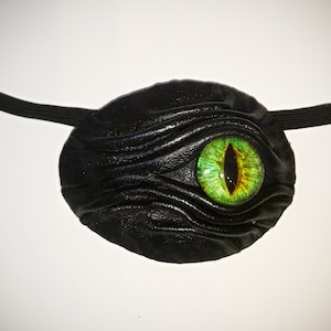May include: A black leather eye patch with a green and yellow eye design.