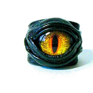 May include: A black leather ring featuring a yellow and orange eye design. The eye is surrounded by black leather that is wrapped around the ring.