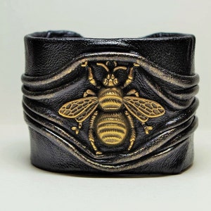 May include: Black leather cuff bracelet with a gold bee design. The bracelet has a textured, wavy design.