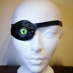 May include: A black leather eye patch with a green eye design. The patch is worn on a white mannequin head.