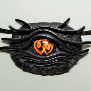 May include: Black leather eye patch with a textured, layered design. The eye patch has a round, orange and yellow glass eye with a swirling pattern.