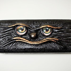Puede incluir: Un estuche rectangular negro para gafas con un diseño de rostro tallado. El estuche presenta dos ojos grandes e iridiscentes, una pequeña nariz y una boca curva, todo delineado en dorado. La superficie texturizada le da al estuche una apariencia artística única.