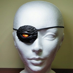 Może przedstawiać: Czarna skórzana opaska na oko z fakturowaną powierzchnią i czerwono-pomarańczowym detalem oka. Opaska na oko jest noszona na białej głowie manekina.