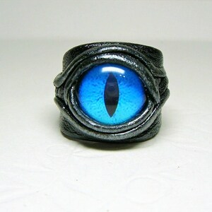 Peut inclure: Une bague en cuir noir avec un motif &oelig;il de chat bleu. La bague est envelopp&eacute;e de cuir et a une finition argent&eacute;e.