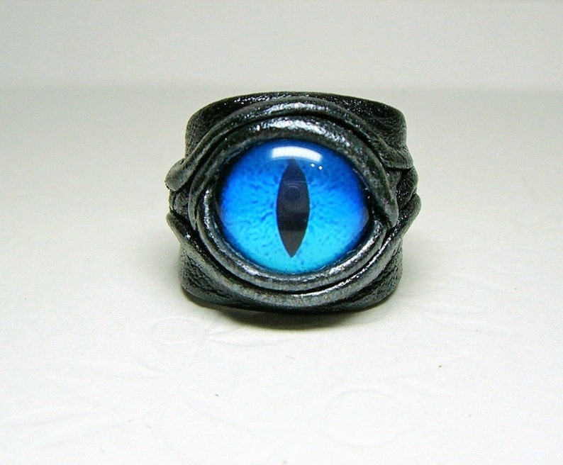 Peut inclure: Une bague en cuir noir avec un motif &oelig;il de chat bleu. La bague est envelopp&eacute;e de cuir et a une finition argent&eacute;e.